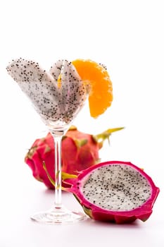 Wedges of a pitaya assorted in a glass and decorated with a mandarine slice. Next to it a halved,and behind it,an entire Fruit..