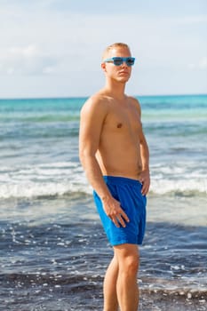 Young man in blue swimming trunks stands on the beach in the blue sea on holiday
