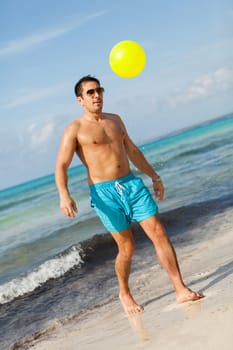 Young attractive man in swim shorts plays football on the beach on holiday in summer