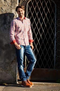 Attractive young man with a plaid shirt and jeans leaning against a wall outdoors in summer