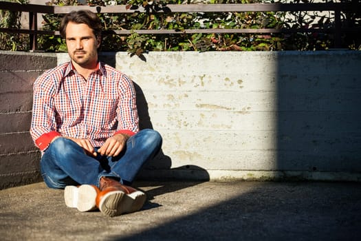 Young man with a checked shirt sits on a wall in the sun and relaxes