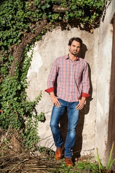 Young man with a plaid shirt and jeans with dark hair and beard outdoors under an efeuranke in daylight