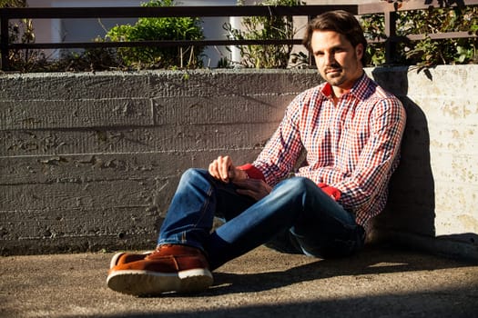Young man with a checked shirt sits on a wall in the sun and relaxes