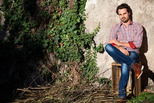 Young man with a plaid shirt and jeans with dark hair and beard outdoors under an efeuranke in daylight