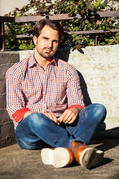 Young man with a checked shirt sits on a wall in the sun and relaxes