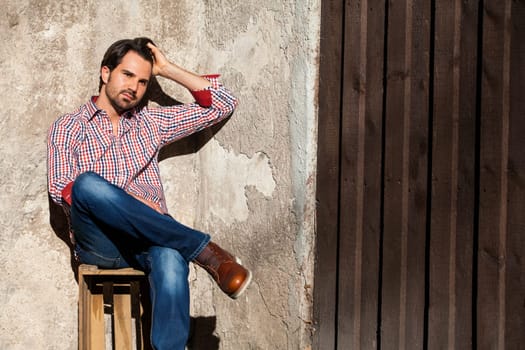 Young man with a plaid shirt and jeans with dark hair and beard outdoors under an efeuranke in daylight