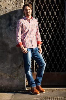 Attractive young man with a plaid shirt and jeans leaning against a wall outdoors in summer