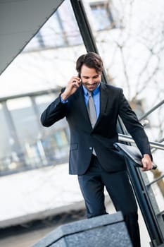 Young trendy successful businessman with mobile phone smartphone cheerful lyrane in front of an outdoor business building