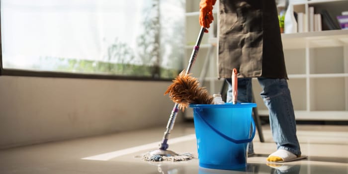 A cleaning woman is standing inside a building holding with a blue tank on the side facilities for tidying up in her hand.