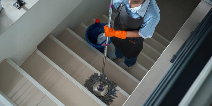 A cleaning woman is standing inside a building holding with a blue tank on the side facilities for tidying up in her hand.