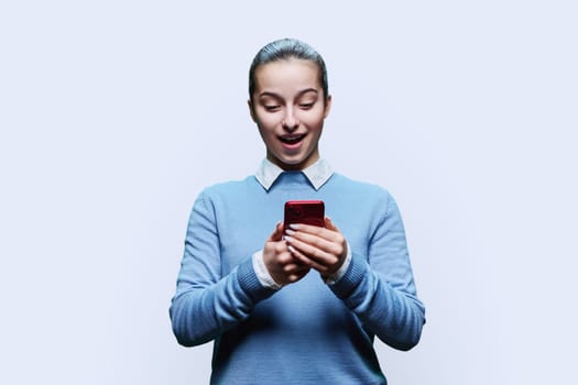 Surprised teenage student, girl 15, 16 years old using smartphone on white studio background. Adolescence, internet technologies, mobile online applications apps services for learning leisure