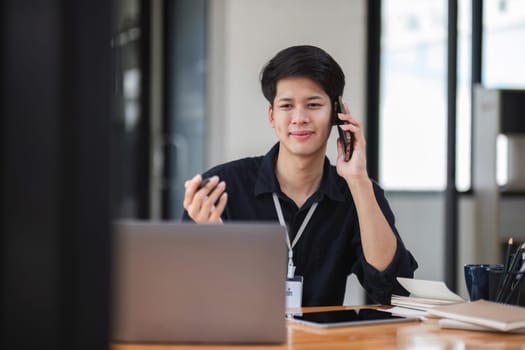 Young businessman talking on the phone with business team using and tablet to do finance, mathematics on table and wood in business office, tax, accounting and analytical research concepts.