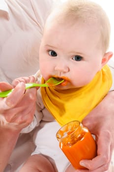 Adult woman sits on a white chair and hades the 6-month-old baby in her arm and fuettes an orange carrot porridge with a yellow plastic loeffel,isolated against a white background.