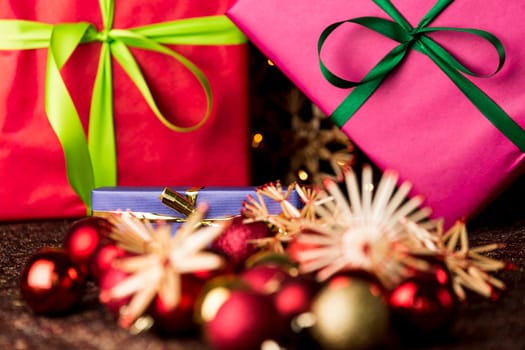 A golden bowknot around a blue gift in the center of the depth of field. Their sharp outlines are contrasting with the blurry forms of baubles and straw stars in the front..