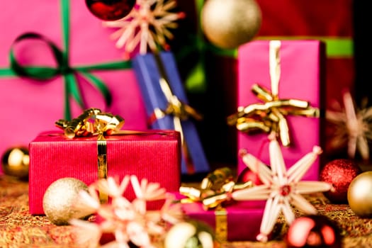 Crimson gift box with golden bow between baubles and straw stars. Five more single-colored packets out of focus. Background for Christmas composition..