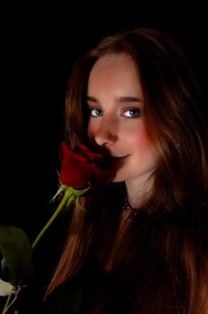A beautiful young woman standing for black background holding a red.rose to her face, looking into the camera..