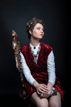Portrait of beautiful Caucasian model in red Victorian-style velvet dress, seated on wooden chair