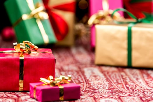 Golden bow knot around a red gift in focus. Blurred shapes of other plain presents in gold,green and magenta outside depth of field. Plenty of text space.