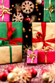 Little magenta gift with golden bow amidst stacked-up Christmas presents. Both wrapping and ribbons are all in red,green and gold. Blurred straw stars and baubles in front and back. Xmas background.
