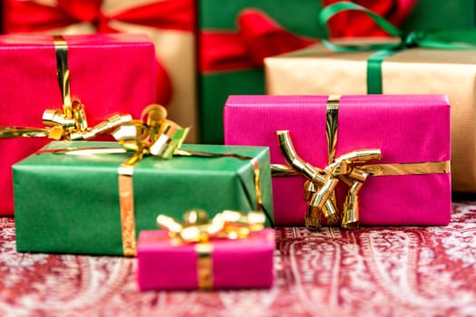 Magenta gift box with golden bow between other plain presents in red, green and gold. Shallow depth of field cuts across the festive cloth. Background for any gift-giving occasion.