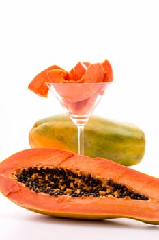 A longitudinal section through the globose body of a papaya fruit. Behind it,there is a cocktail glass filled with peeled papaya slices and an entire fruit. The focus is on the halved fruit in front..