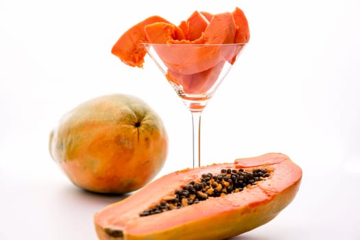 Blazing tangerine slices of the papaw fruit assorted in a long-stemmed glass. A papaw halved along its longitudinal axis in the foreground and a whole fruit in the back..