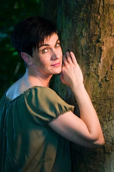 Head-and-shoulder portrait of a middle-aged black-haired woman leaning against a tree in the narrow beam of light from the top right,friendly looking into the camera