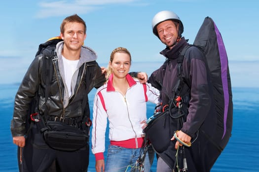 Men, woman and portrait with happy for paragliding, extreme sport and outdoor by sea in sunshine. Group, fitness and smile on adventure with helmet, backpack or parachute for jump, safety or blue sky.
