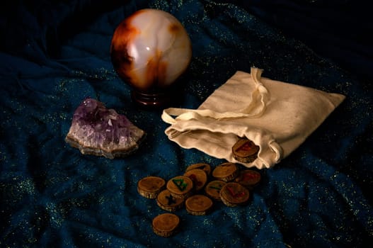 A plain muslin bag with wooden runes spilling out over a sparkling blue cloth with amethyst and crystal ball in the background.