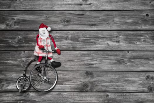 Funny red santa claus on wooden grey background in hurry for buying christmas presents sitting on a old bike.
