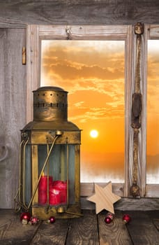 Christmas background with red candle and lantern - in the back a sunset. Windowsill view in orange colors.
