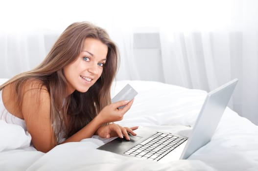 Young woman using laptop in bed with credit card