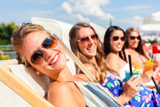 Four women drink cocktails in a beach club and lie on a sun lounger