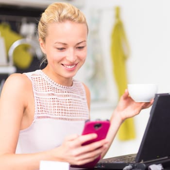 Business woman working from her home. Multitasking. Having morning coffee while texting on the smarth phone and working on leptop computer.