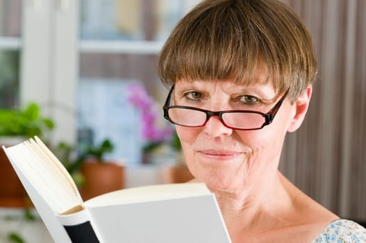 Inside shot head-and-shoulder portrait of an older woman with reading glasses and open book friendly smiling into the camera
