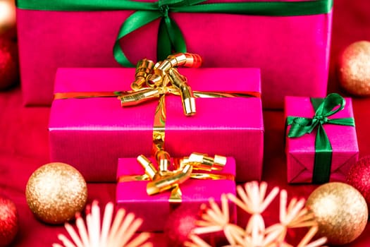 Four magenta gifts ready for handing out of presents. Surrounded by blurred Christmas bulbs and straw stars on a red cloth. Narrow depth of field runs across the golden bow of the medium-sized box.
