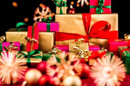 Heap of Christmas gifts in mainly golden and red colors. Blurred baubles and stars on red blanket in front. Bokeh effect against pitch black background. Shallow depth of field.