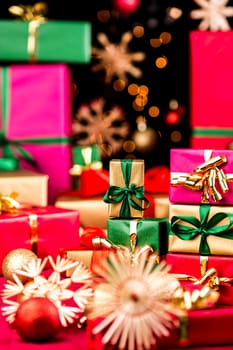 Small, single-colored Christmas presents piled up between stars and baubles. Narrow depth of field. Focus on small golden box with green bow. Out-of-focus points of light against black background.