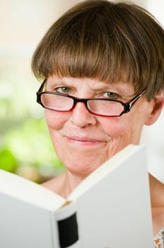 Inside shot head-and-shoulder portrait of an older woman with reading glasses and open book friendly smiling into the camera