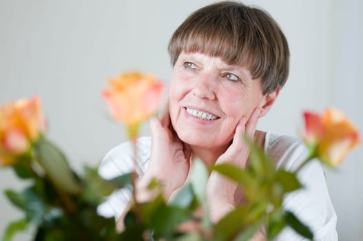 Frontal portrait of an elderly woman sitting behind flowers at the table holding her hands to her cheeks and looking obliquely up left