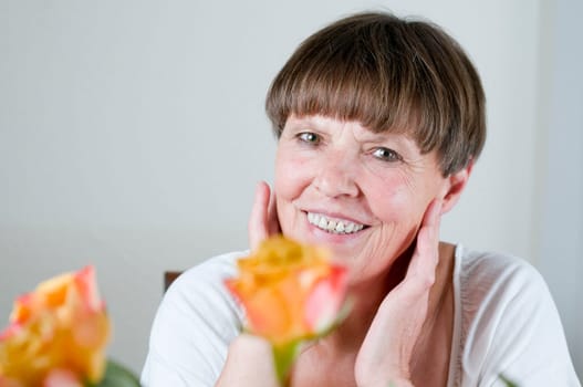 Frontal portrait of an elderly woman sitting behind flowers at the table holding her hands to her cheeks and smiling at the camera