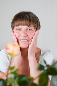Frontal portrait of an elderly woman sitting behind flowers at the table holding her hands to her cheeks and smiling at the camera