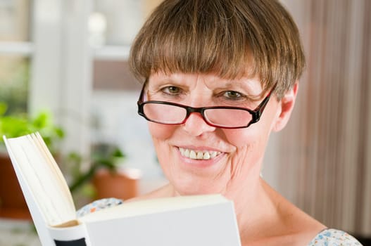 Inside shot head-and-shoulder portrait of an older woman with reading glasses and open book friendly smiling into the camera