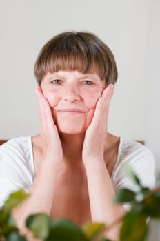 Frontal portrait of an elderly woman sitting behind flowers at the table holding her hands to her cheeks and smiling at the camera