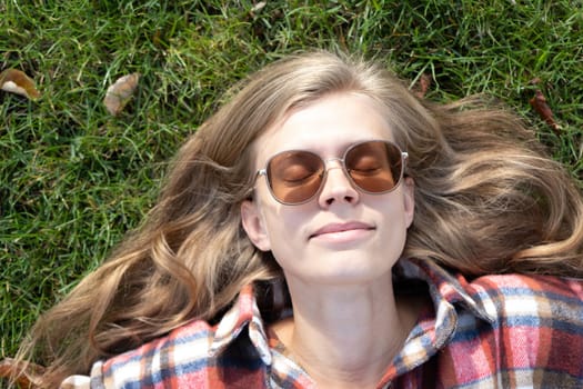 young caucasian woman in red plaid shirt smiling while relaxing on green grass with yellow autumn leaves