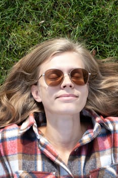 young caucasian woman in red plaid shirt smiling while relaxing on green grass with yellow autumn leaves