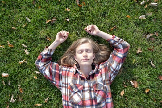 young caucasian woman in red plaid shirt smiling while relaxing on green grass with yellow autumn leaves