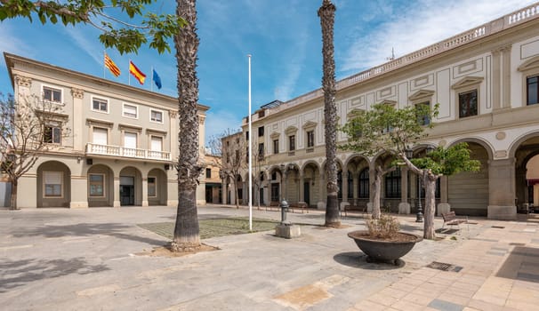 Town hall with flags on roof and paved square in Vilassar de Mar on summer day. Architectural landmarks of Catalonia attracting tourists at noon