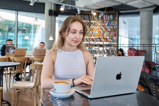 Warsaw, Poland, 12.05.2023. Beautiful girl inside cafe with cup of coffee using laptop. Attractive young blonde woman with curly hair working studying in cafe. Beauty, youth, lifestyle concept