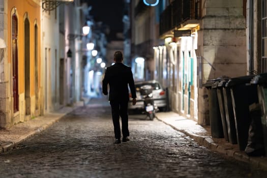 A solitary figure traverses an old street, buildings bathed in warm night lights - view from behind